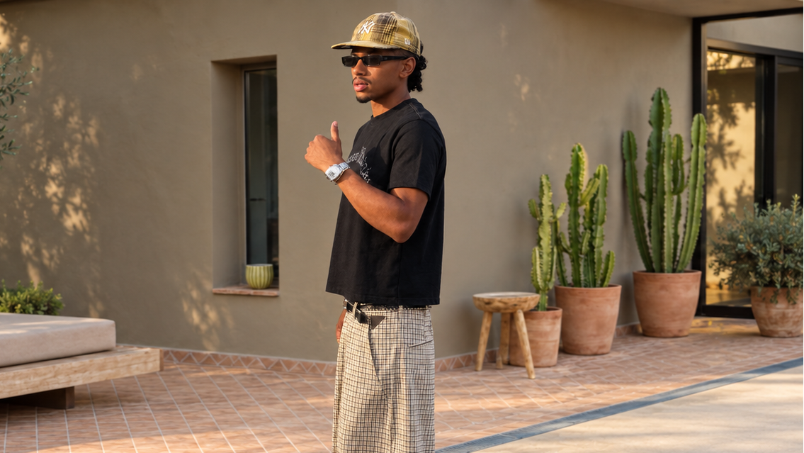 A man wearing a black t-shirt, beige and white checkered shorts, a beige cap, and red flip-flops stands on a tiled patio in front of a stucco building with a daybed and potted cacti.